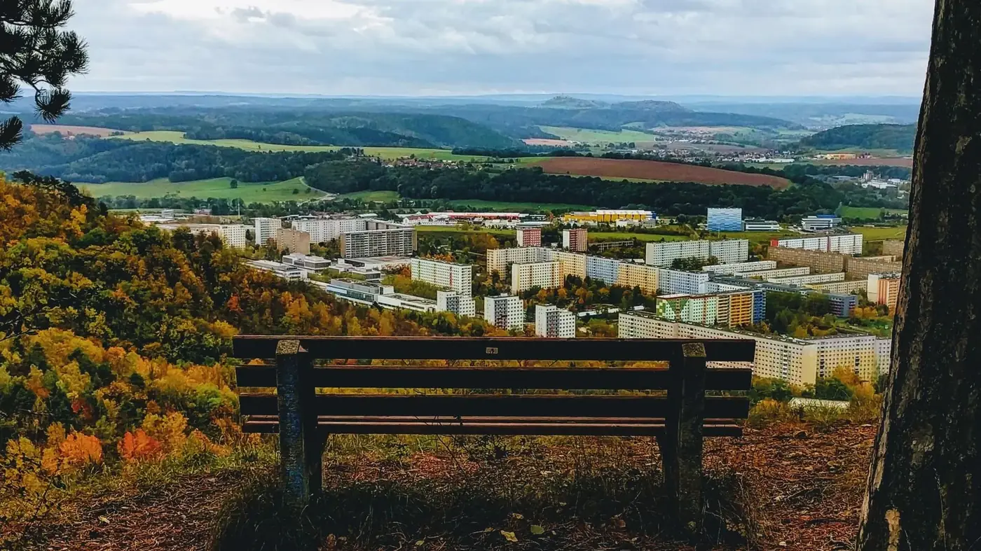 A bench overlooks a cityscape nestled in rolling hills with colorful autumn trees.