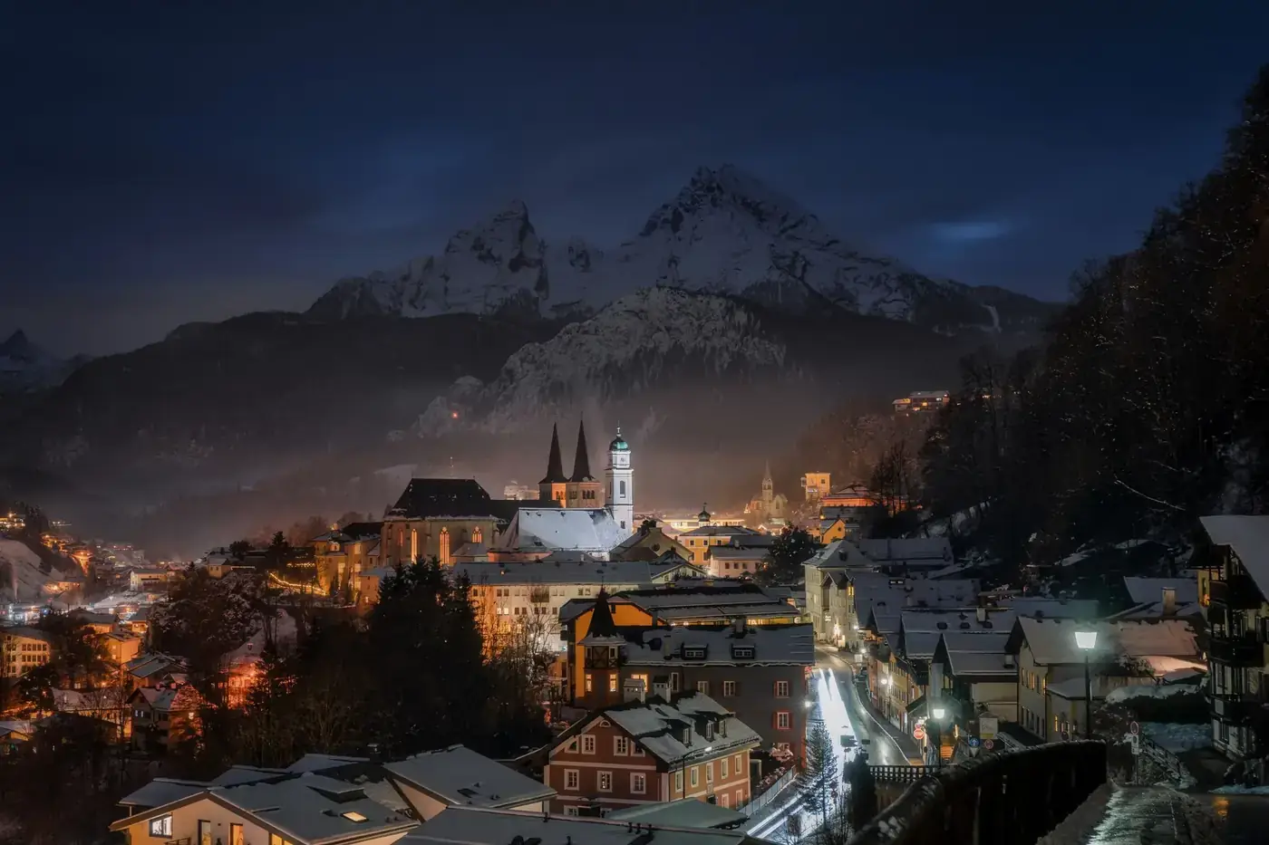 A picturesque winter night view of Berchtesgaden, Germany, with snow-covered rooftops and majestic mountains in the back