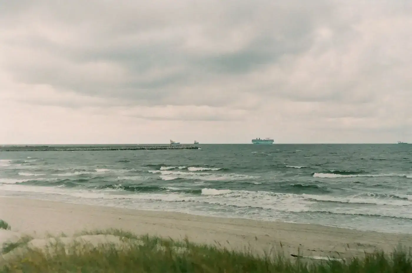 Calm ocean waves on a sandy beach with ships in the distance under a cloudy sky.