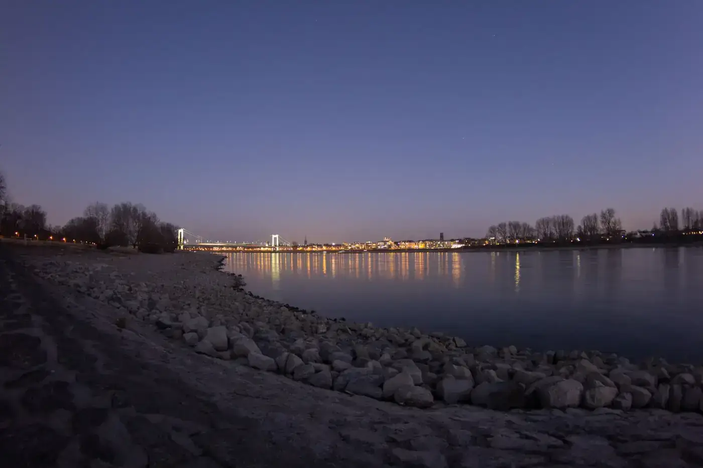 A serene twilight scene along the Rhine River in Cologne, showcasing city lights reflecting on calm waters.