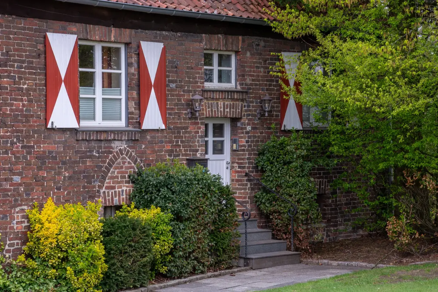A charming brick house with white and red shutters, surrounded by lush greenery in Coesfeld, Germany.