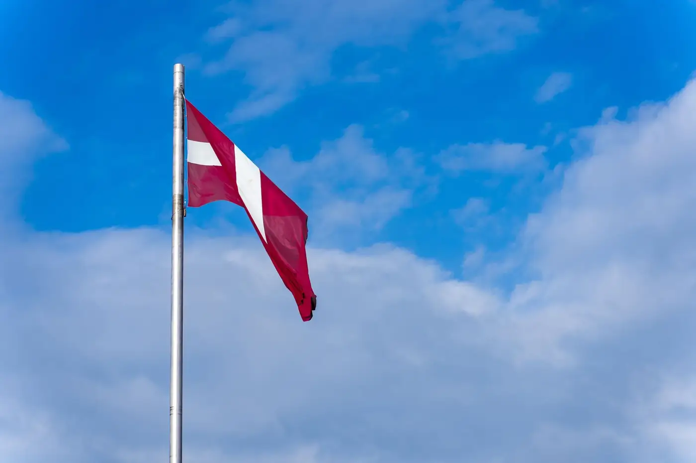 A Latvian flag on a flagpole waving against a backdrop of clear blue sky and clouds.