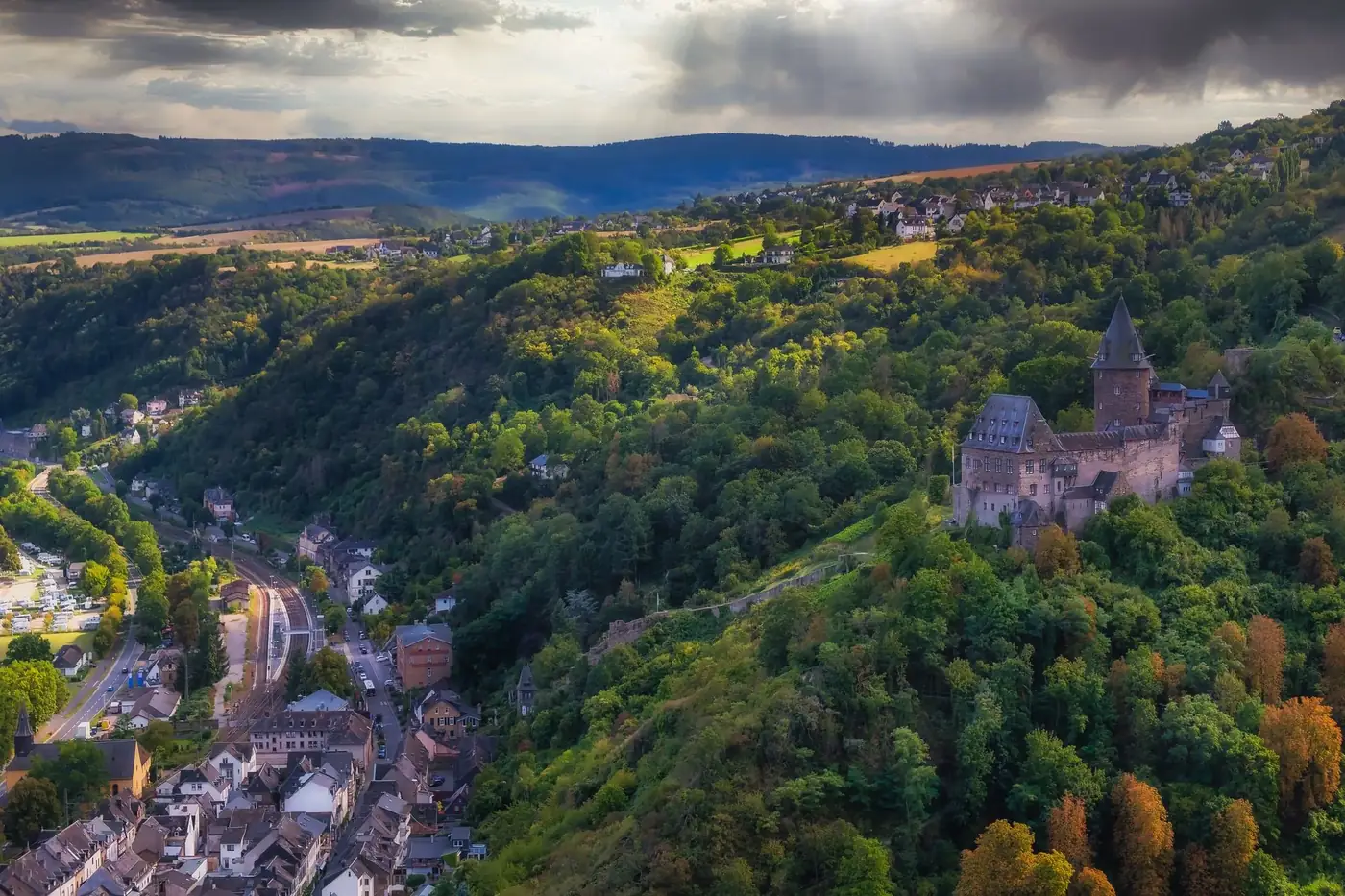 Scenic aerial view of a medieval castle on a hill surrounded by a lush forest and a quaint town.