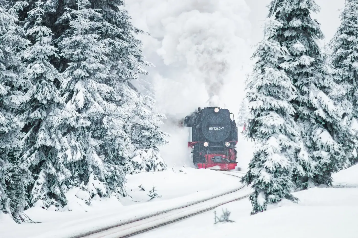 A steam train moves through a snowy forest landscape, surrounded by frosted trees in winter.