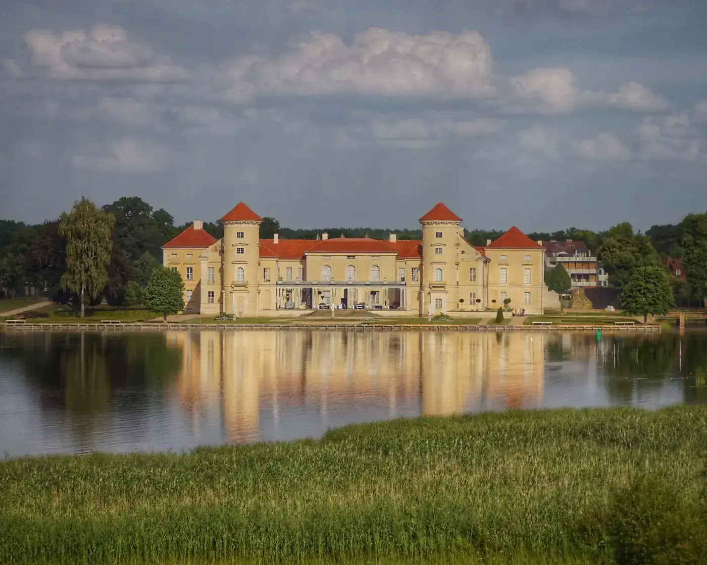 Scenic view of a historic castle with red roofs reflecting in a still lake under a blue sky.