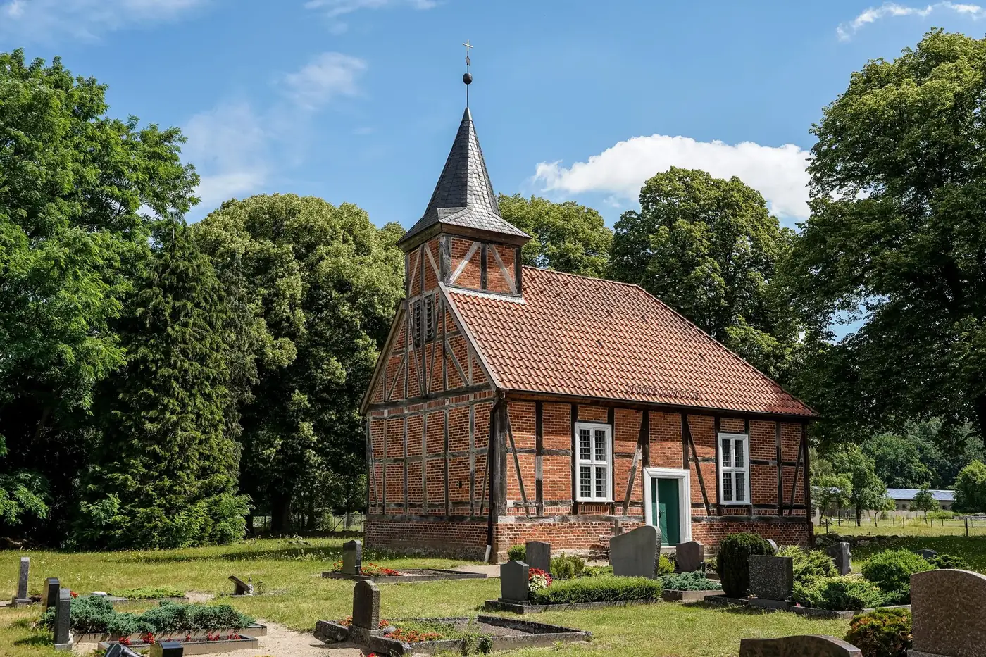 Charming 18th-century brick and timber church in Greven, surrounded by lush greenery and gravestones.