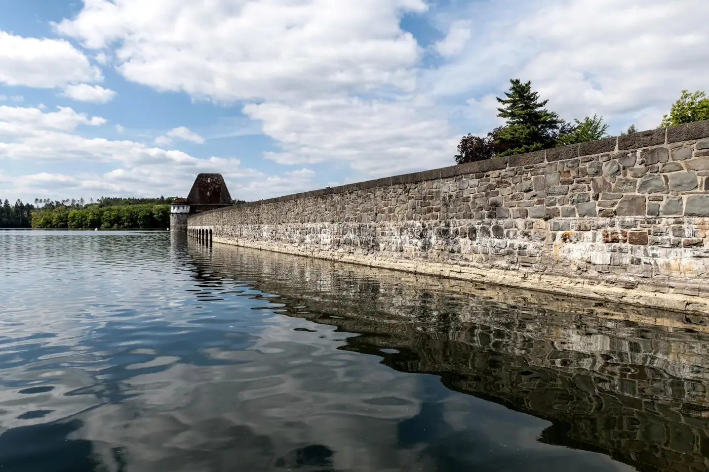 Serene view of the Möhne Reservoir dam wall, surrounded by nature under a partly cloudy sky.