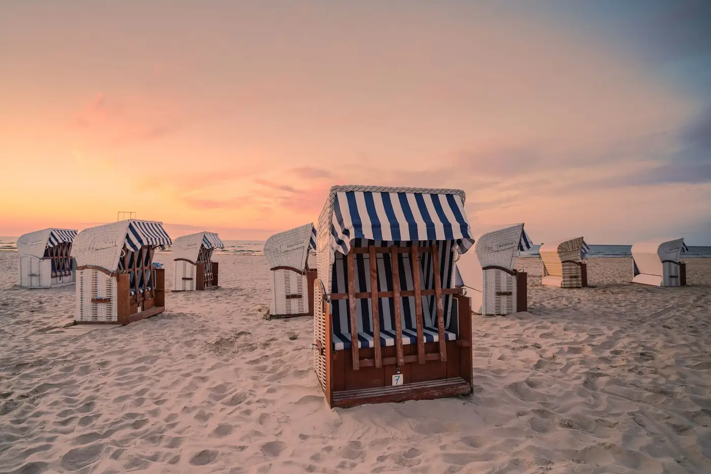 Striped beach cabins line a sandy shore at sunset in Świnoujście, Poland, offering a serene coastal scene.