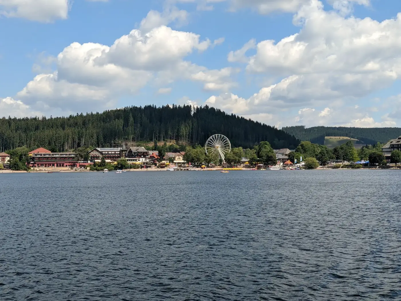A beautiful lakeside view featuring a ferris wheel and a small town, surrounded by lush green hills under a blue sky.
