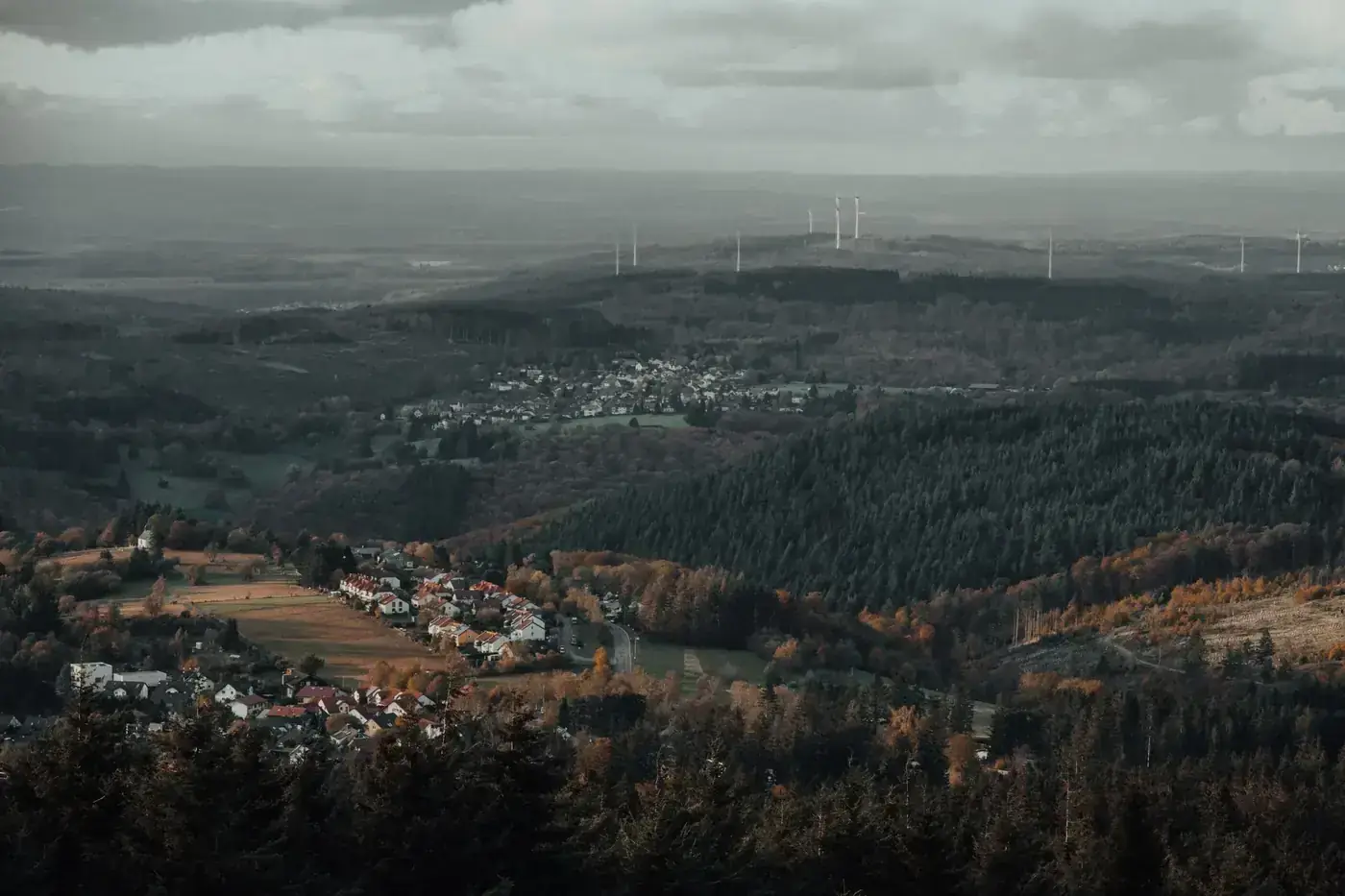 A scenic aerial view of a forested valley with a town and wind turbines during sunset.