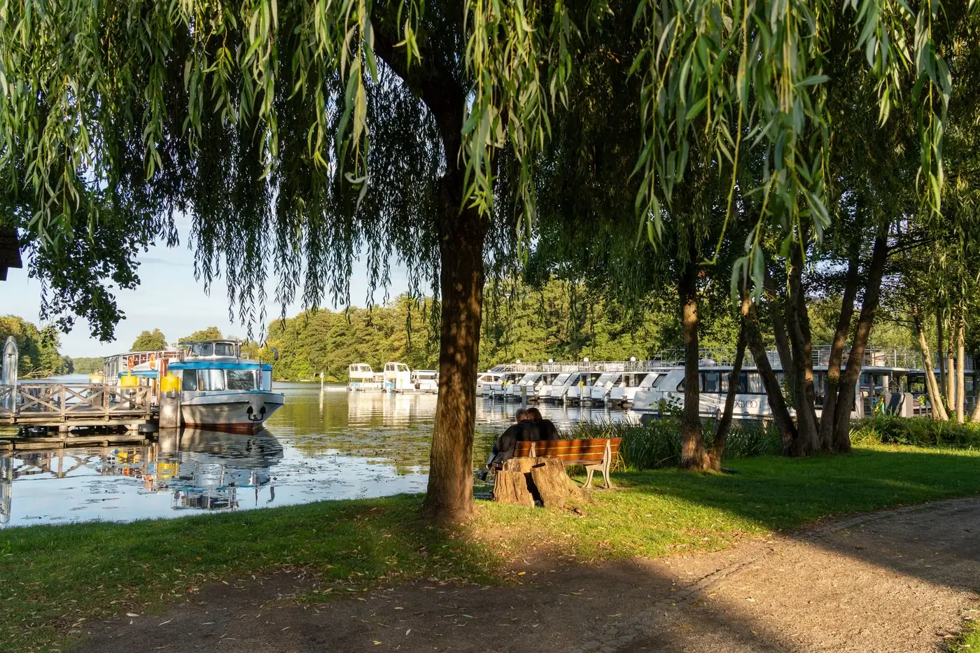 Idyllic lakeside scene with boats, willow tree, and peaceful marina in summer sunlight.