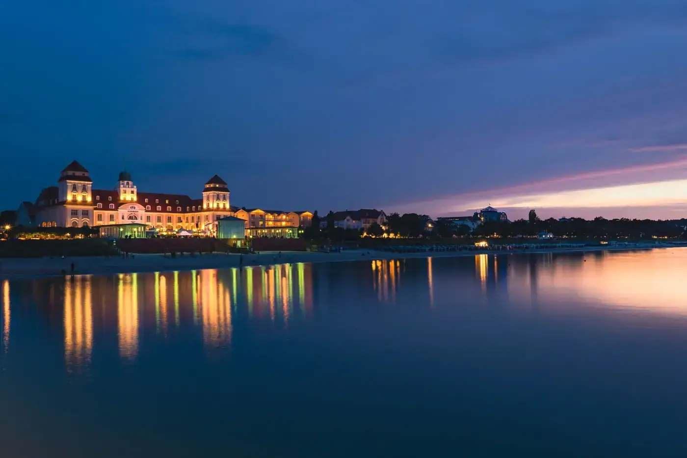 Beautifully illuminated castle-like building reflecting on water during twilight, set against a serene backdrop.