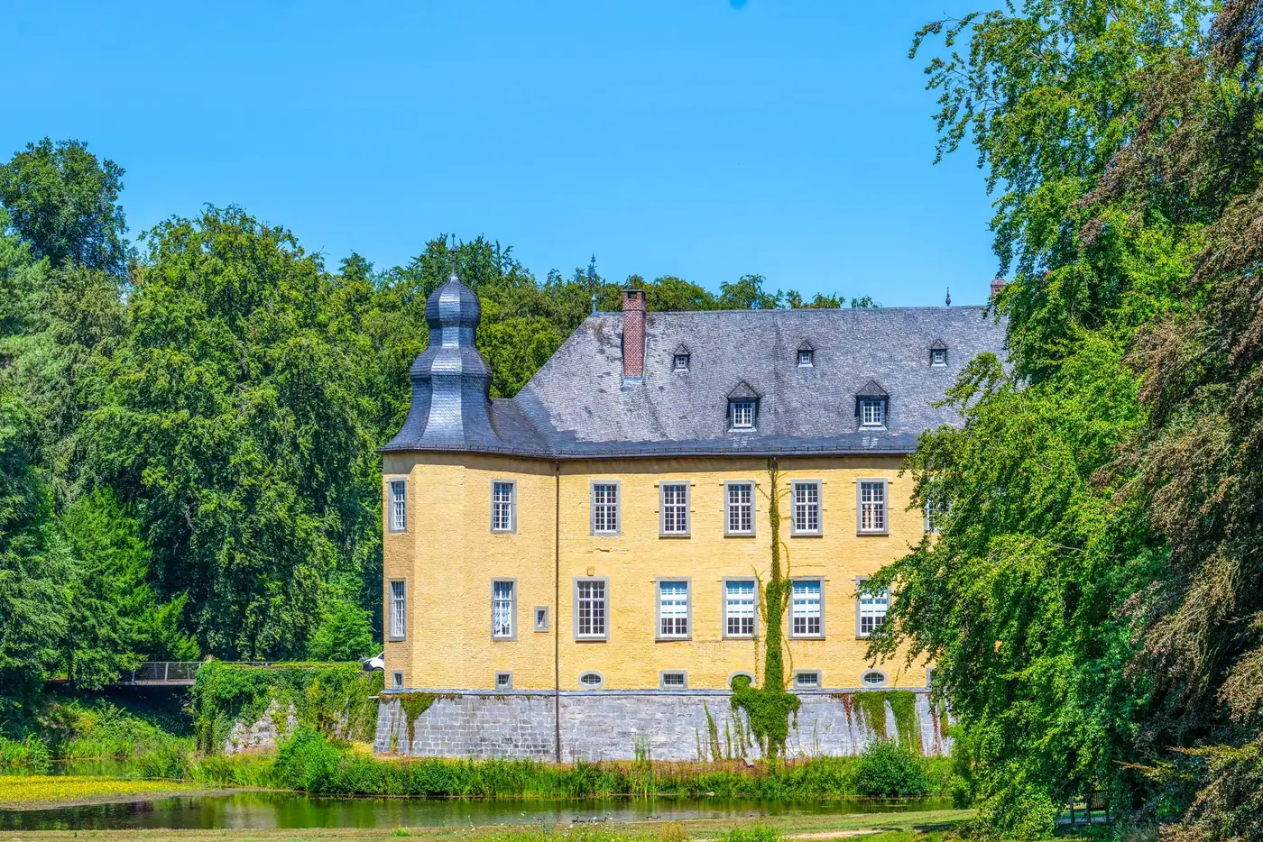 Scenic view of a historic yellow castle surrounded by lush greenery and a pond.